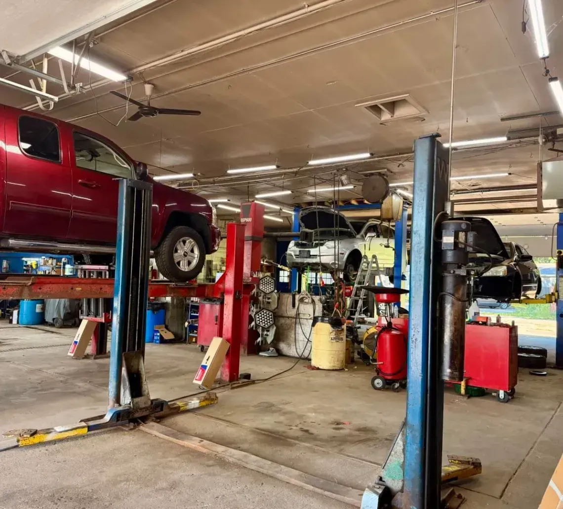 A repair shop with a red truck on a lift, a car with its hood open in the background, and various automotive equipment.