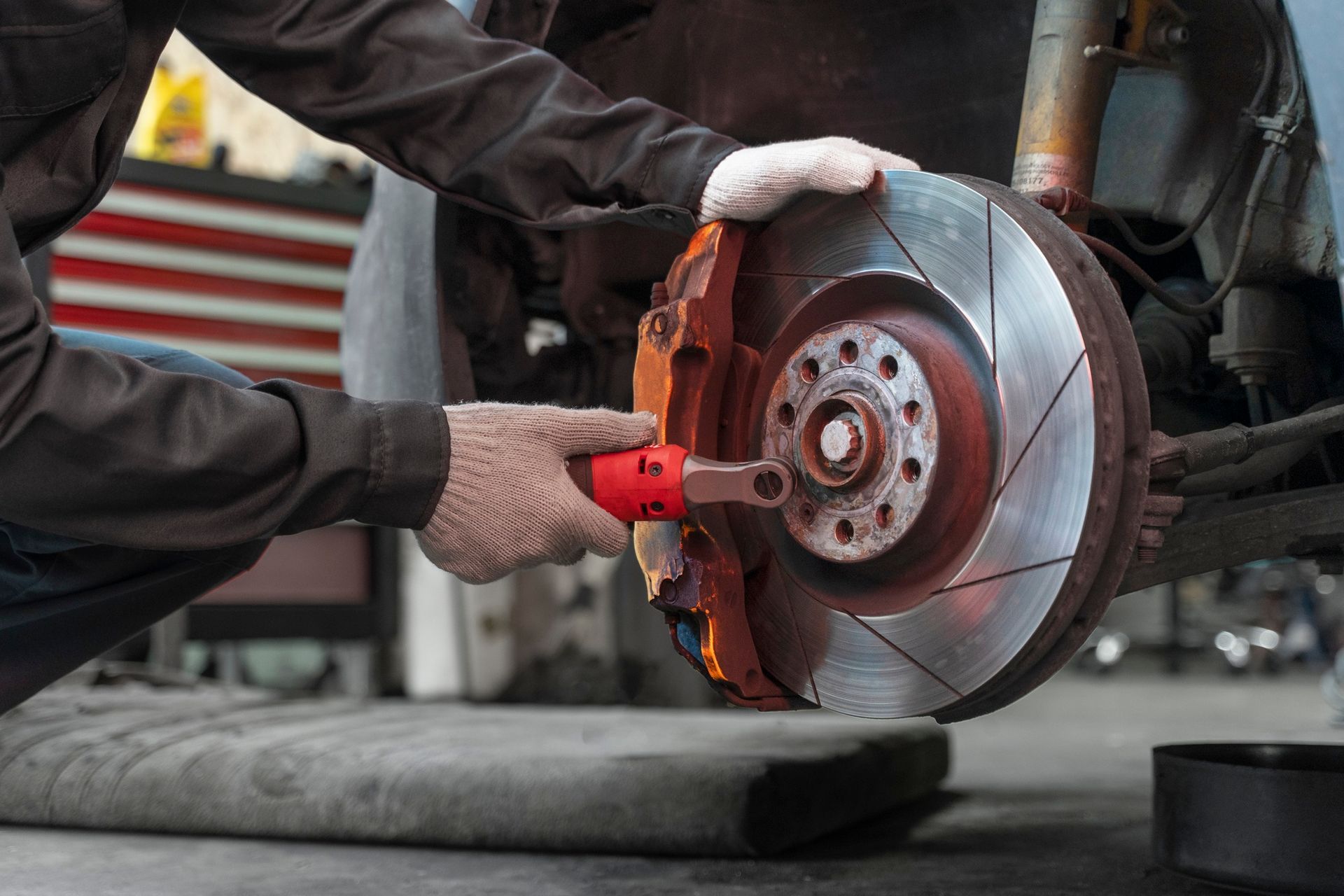 A mechanic in a workshop uses a red tool to tighten a bolt on a car's slotted brake disc rotor.