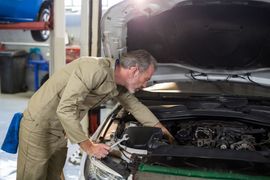 A mechanic in beige coveralls uses a wrench to work on the engine of a car with its hood open in a repair shop.