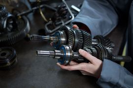 A mechanic's hands holding metal gear assemblies from a vehicle transmission in a workshop.