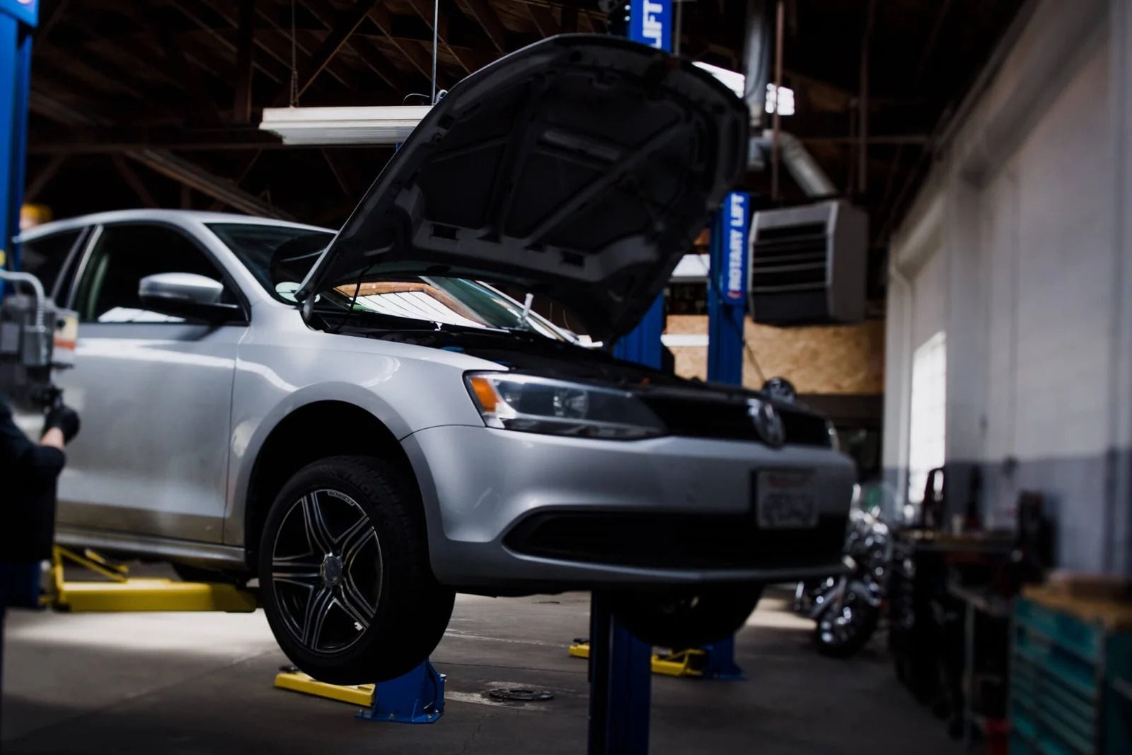 A silver sedan with its hood open, elevated on a hydraulic lift inside an auto repair shop.