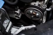 A close-up view of an automotive engine component, featuring a pulley wheel, drive belt, and metallic mounting brackets.