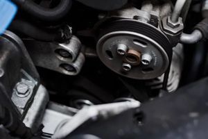 A close-up view of an automotive engine component, featuring a pulley wheel, drive belt, and metallic mounting brackets.