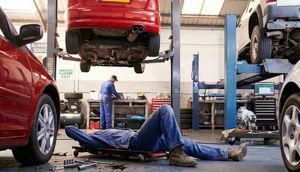 Two mechanics working in an auto repair shop; one lies on a creeper under a car while another stands at a workbench.