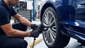 A mechanic wearing black gloves uses a pneumatic impact wrench to tighten a bolt on a car wheel in an auto repair shop.