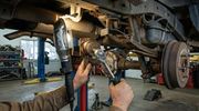 A mechanic uses a ratchet to work on the rear axle and suspension of a vehicle raised on a lift in an auto repair shop.