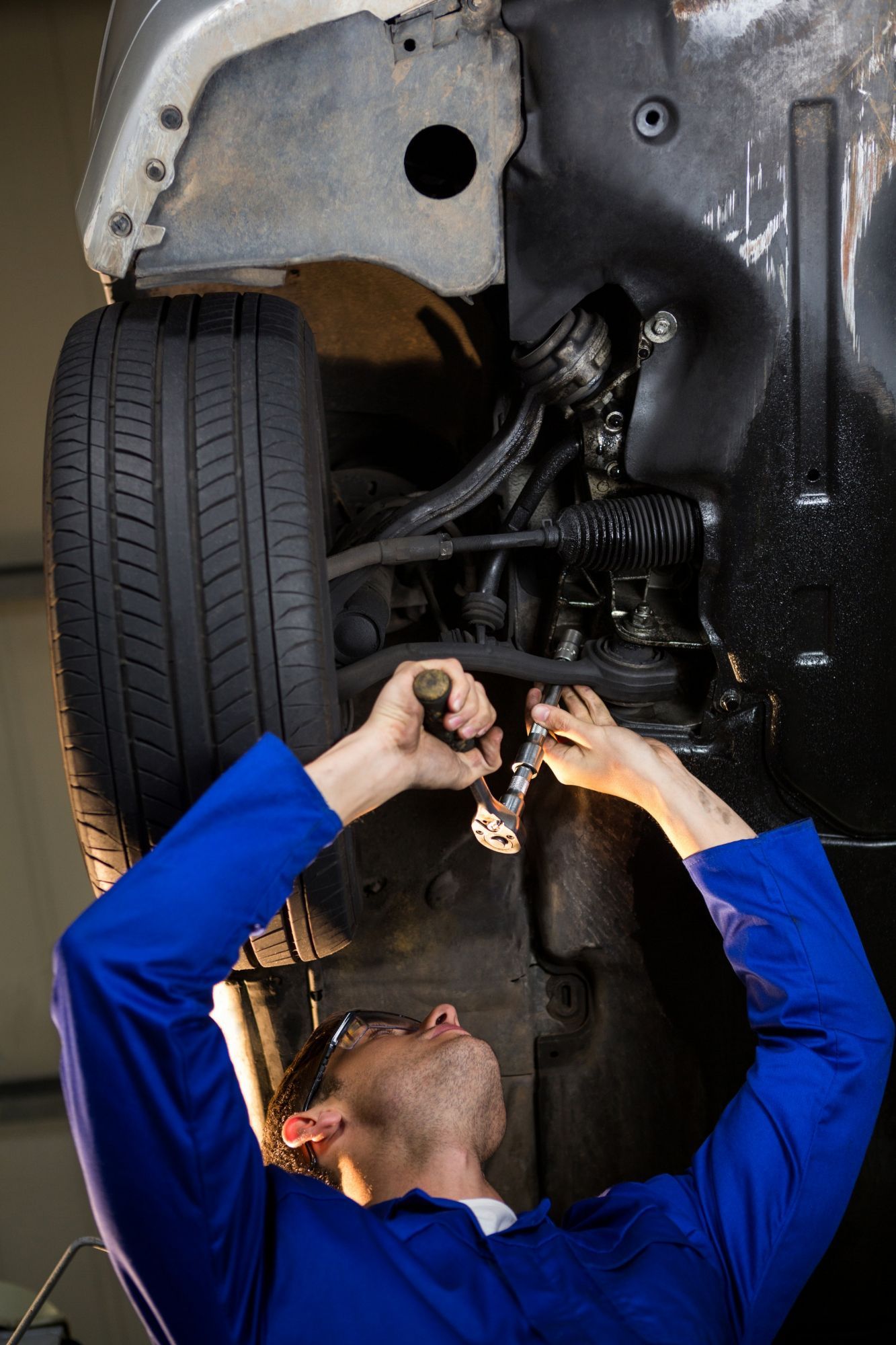A mechanic wearing blue coveralls uses a wrench to work on the underside of a car raised on a lift in a garage.