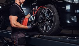 A mechanic uses a red tool to adjust a black car tire on a lift in a workshop.
