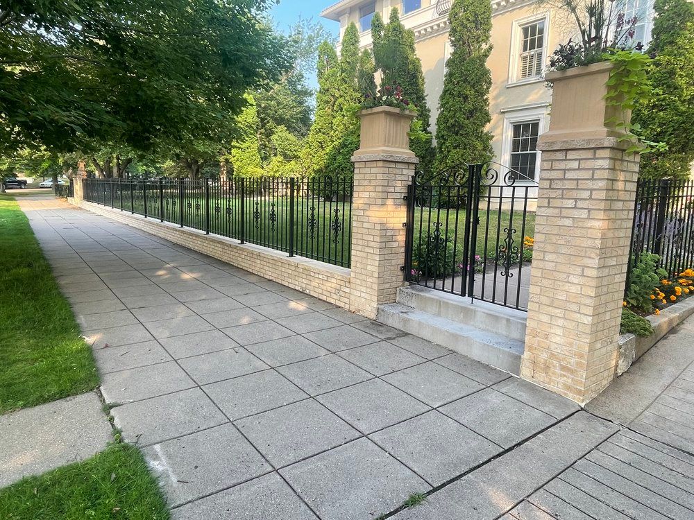 A brick walkway leading to a large house with a wrought iron fence.