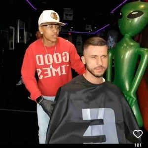 A man is getting his hair cut by a man wearing a red shirt that says good eimi