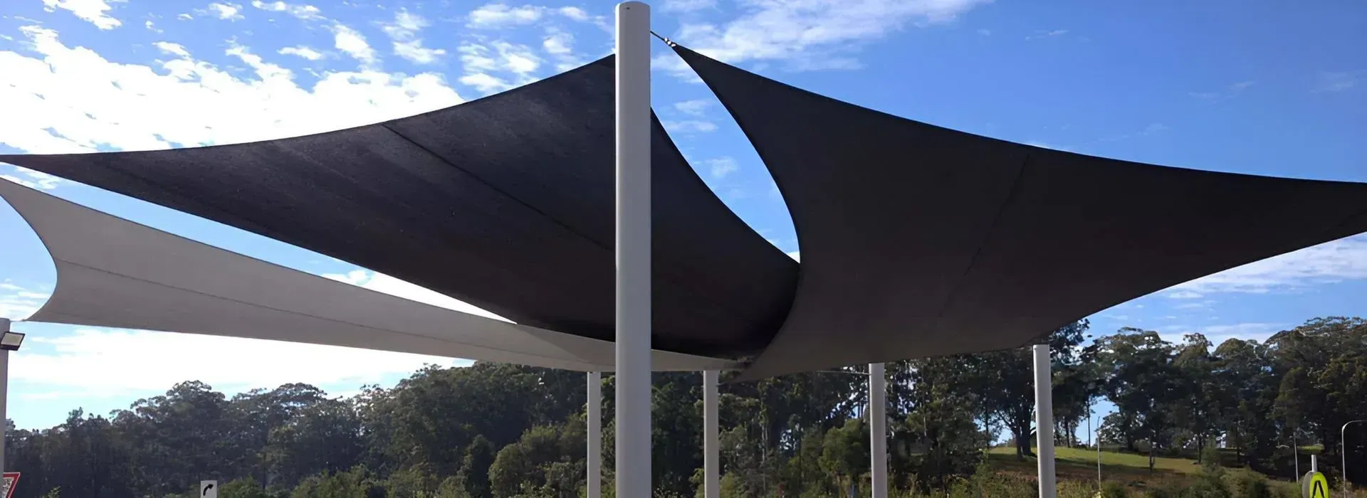 Black and white shade sails stretched between tall poles under blue sky, with lush green trees.