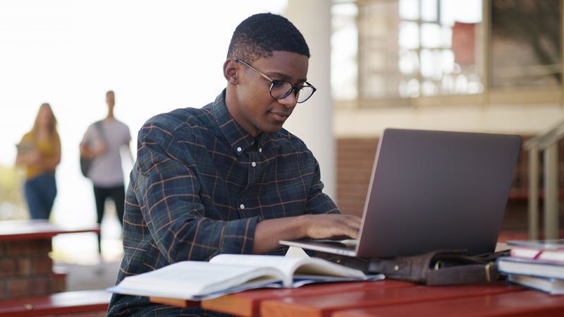 A student wearing glasses works on a laptop, sending college fair follow-up emails.