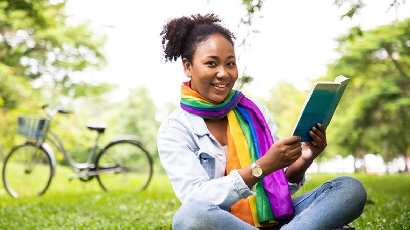 A person wearing a rainbow scarf smiles a book, sitting on the grass in a park with a bicycle nearby.