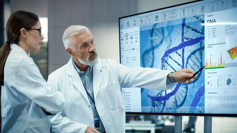Two scientists in white lab coats review DNA data displayed on a large digital screen in a laboratory.