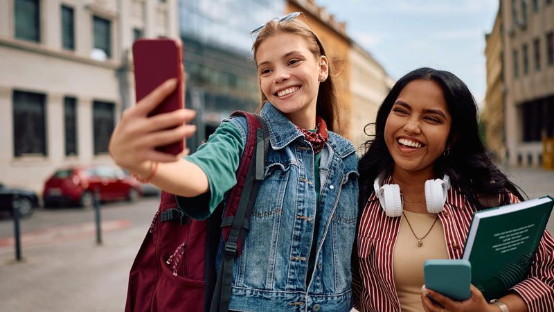 Two students smiling while taking a selfie together on a city street, one holding a phone and the other carrying a book.