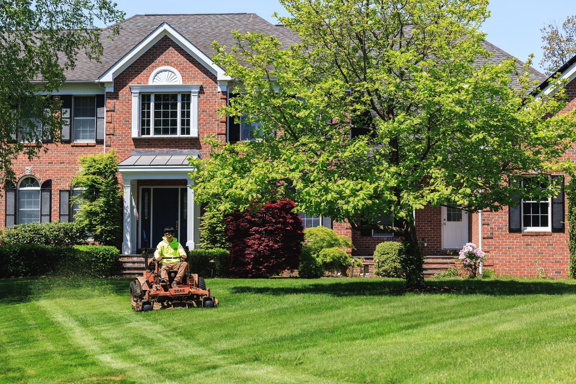 Person mowing a lawn in front of a brick house with lush green grass and trees.