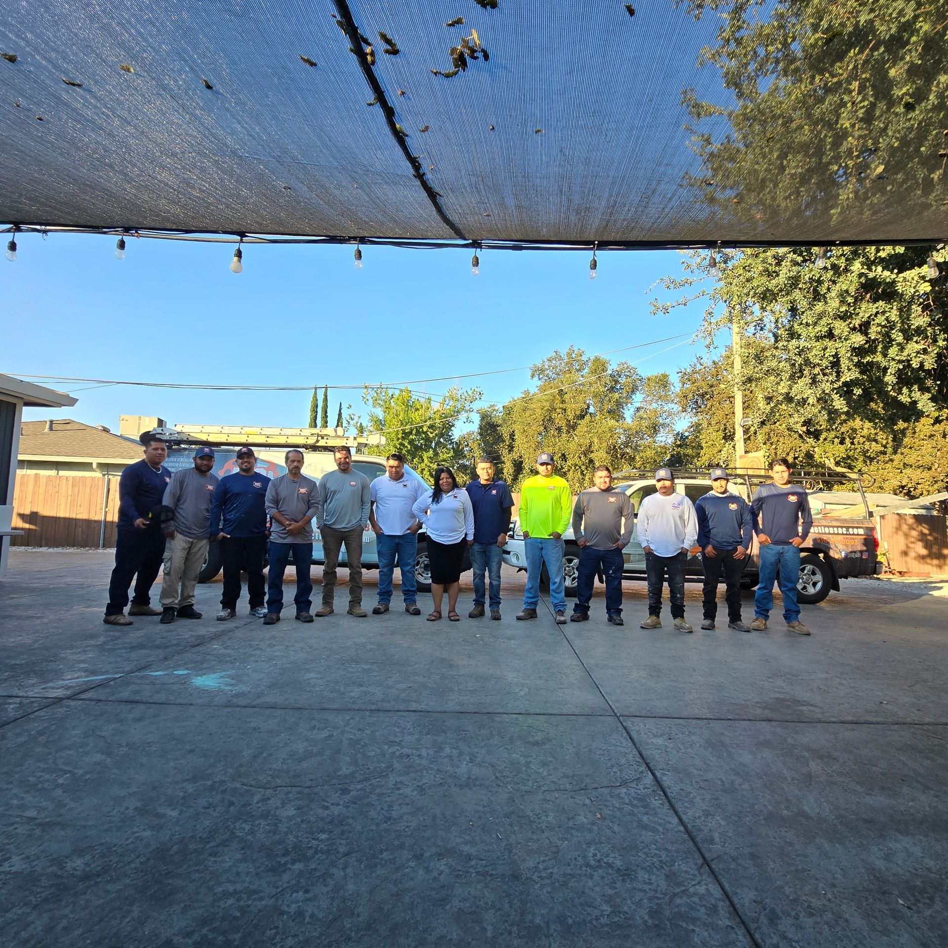 Group of people standing outdoors in a parking lot under a shade canopy, facing the camera.