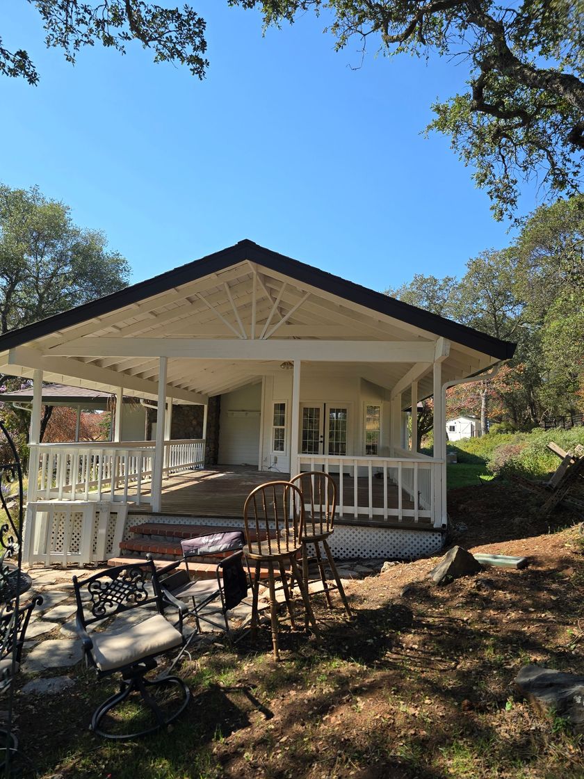 White porch-covered cottage with chairs and stone patio under trees on a sunny day