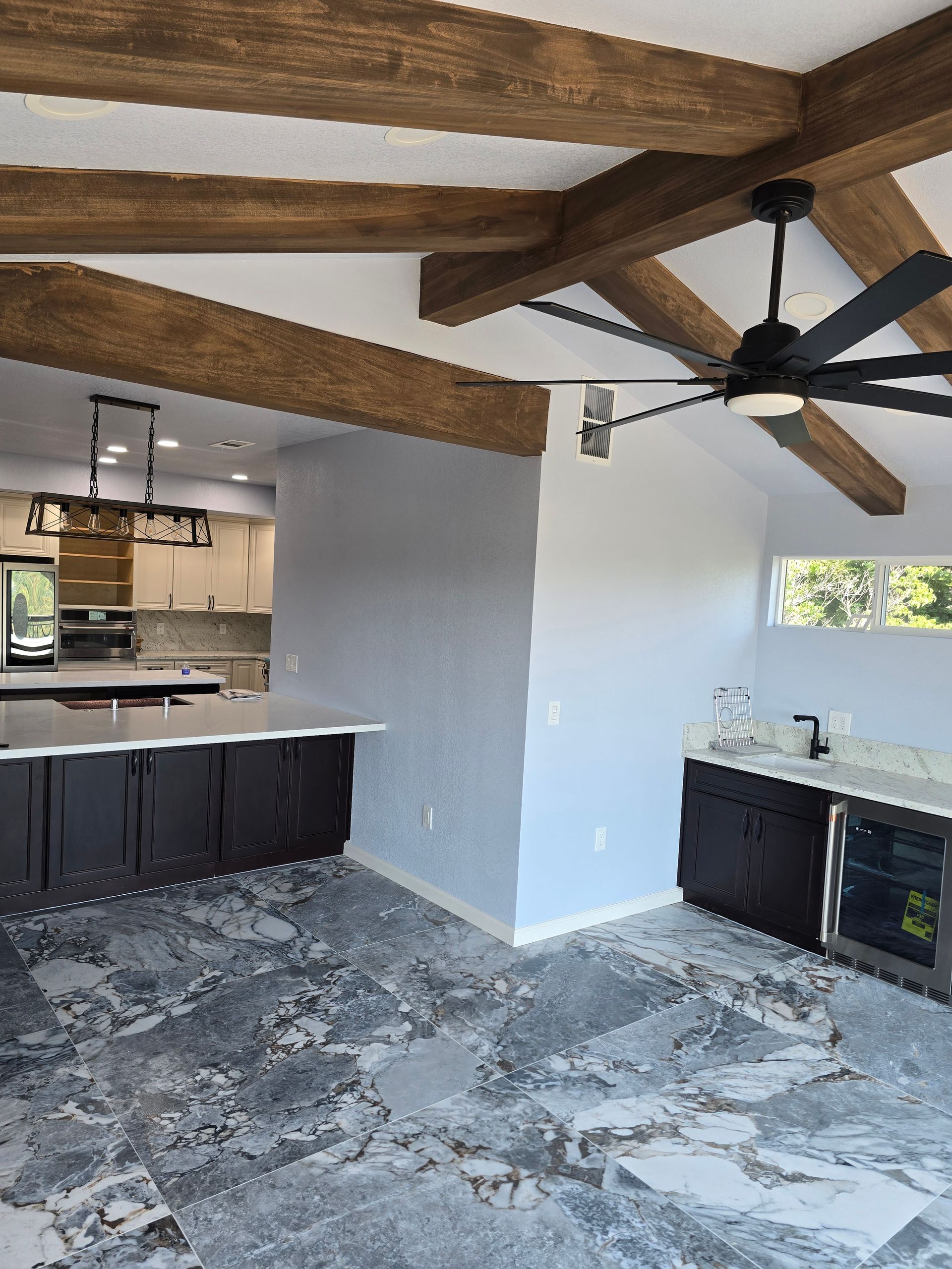 Open kitchen and living area with exposed wood beams, dark cabinets, ceiling fan, and stone-pattern tile floor