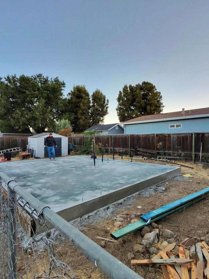Concrete foundation under construction beside a house, with workers, tools, and gravel on site.