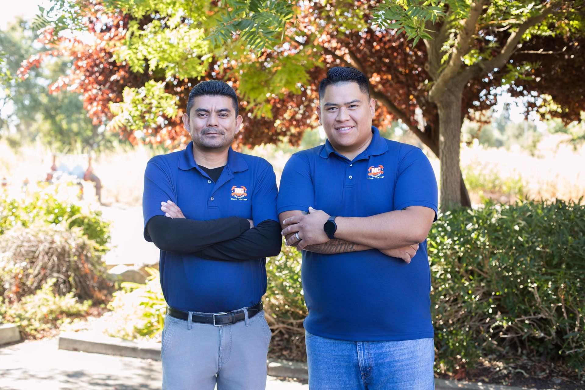 Two men in blue polos stand with arms crossed outdoors under a tree.