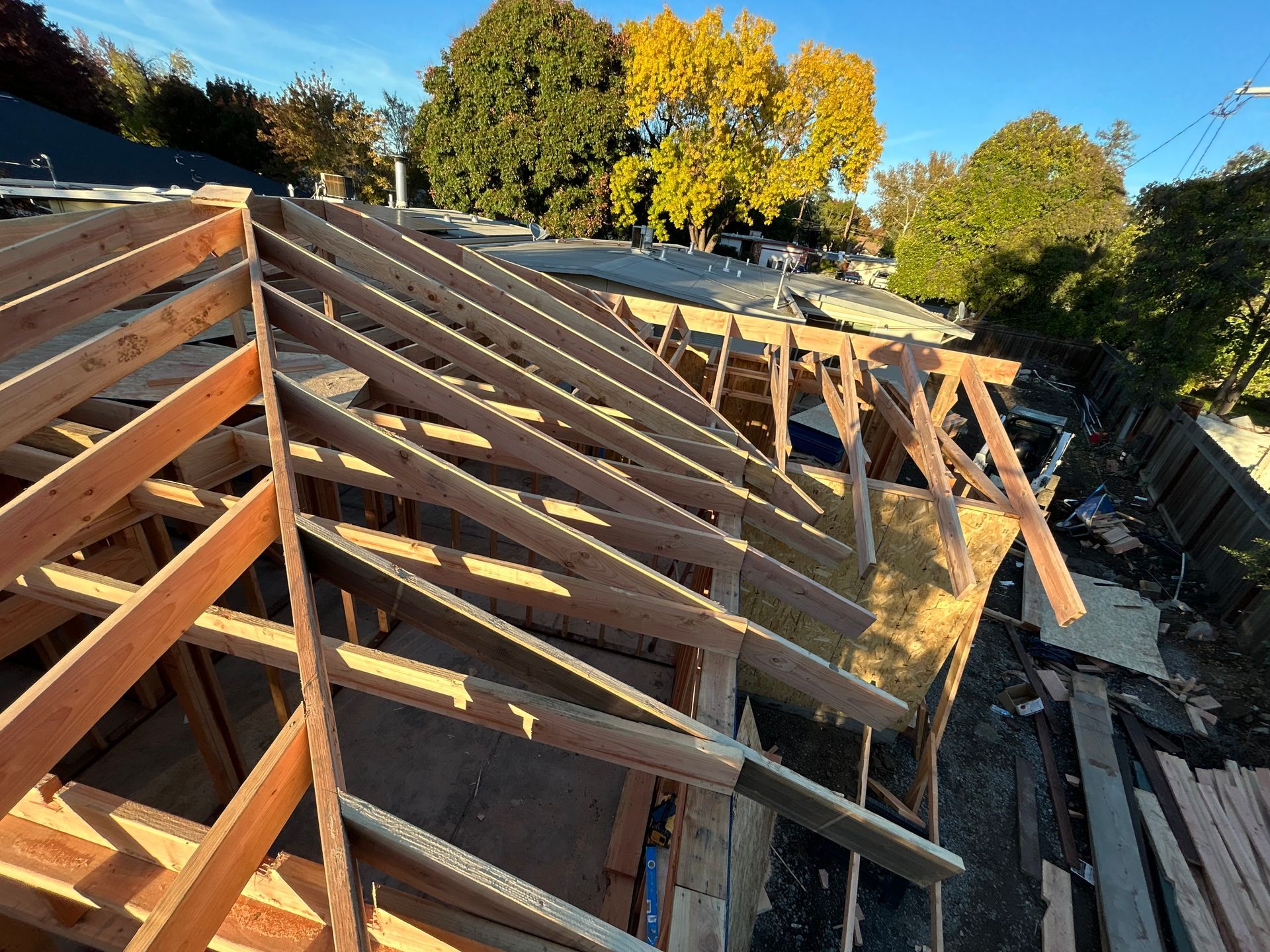 Collapsed wooden roof framing on a damaged house, with debris and autumn trees in the background