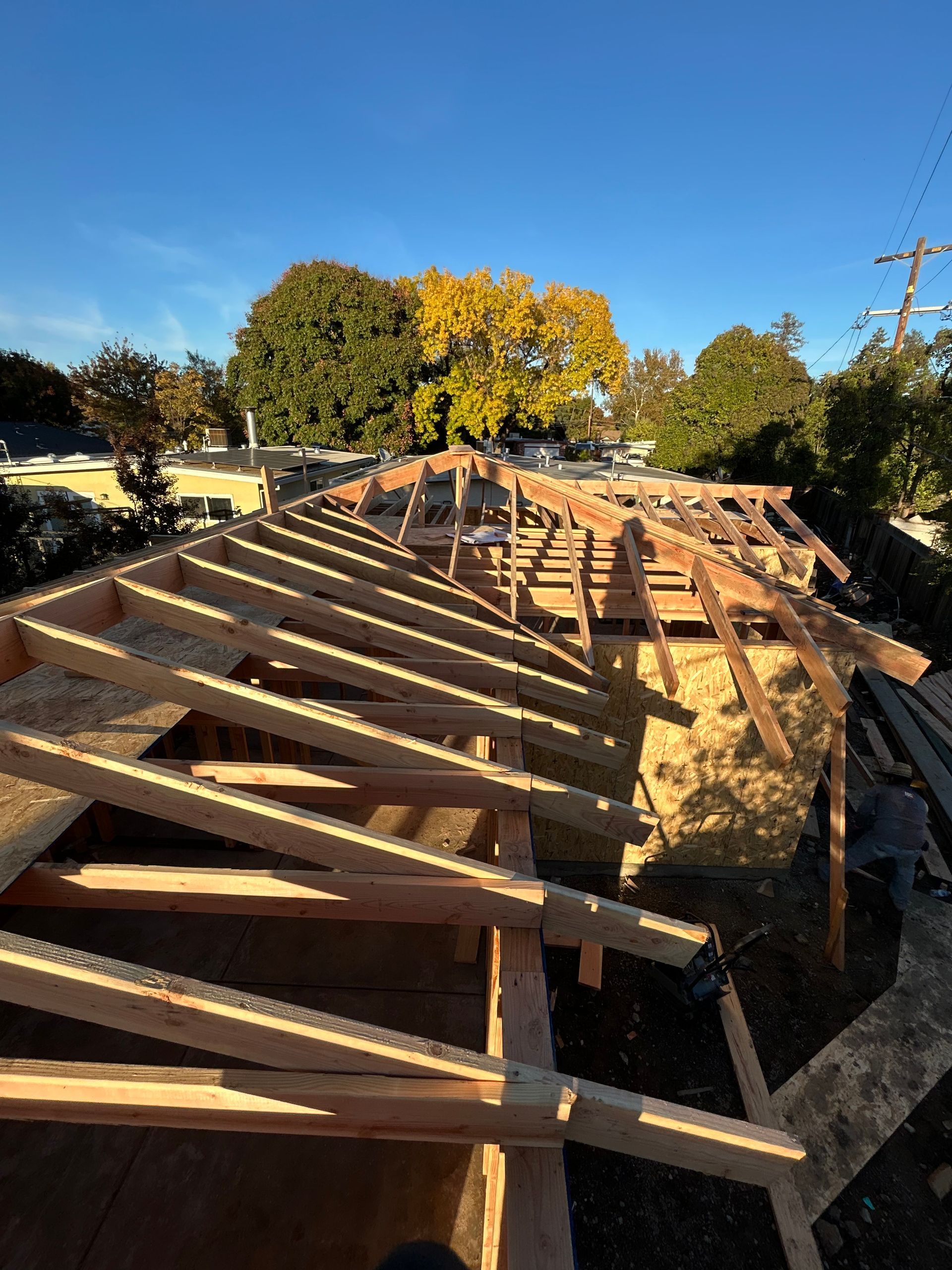Wooden roof framing under construction beside a trench, with autumn trees and blue sky.