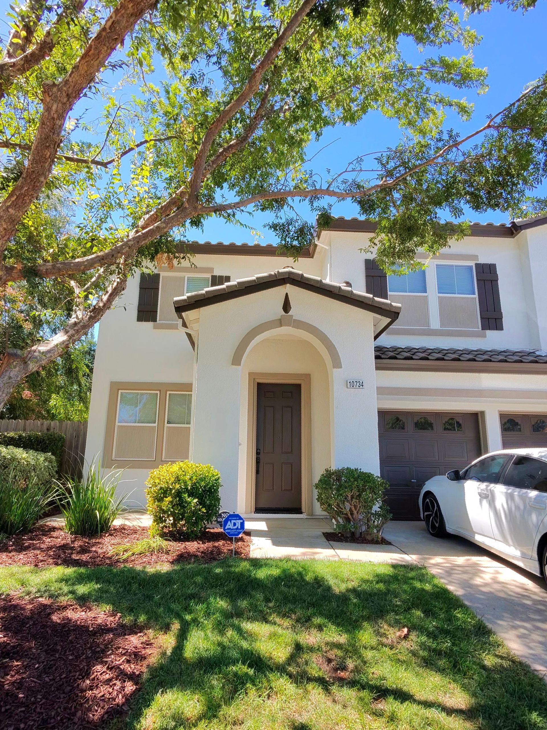 Sunny suburban house exterior with arched entry, tree shade, and a white car in the driveway