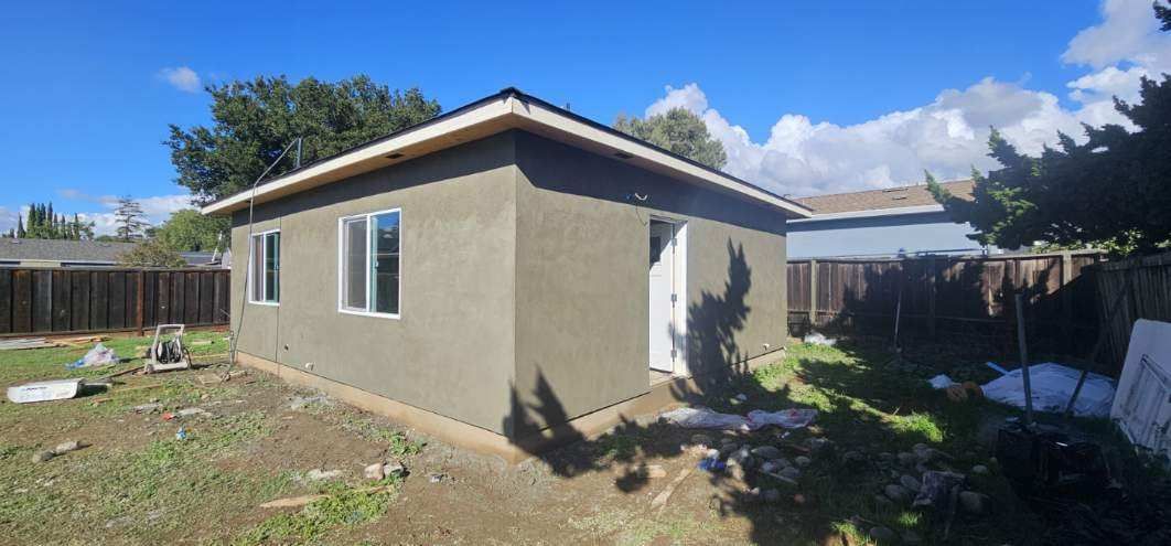 Small tan house with white trim in a backyard under a blue sky.