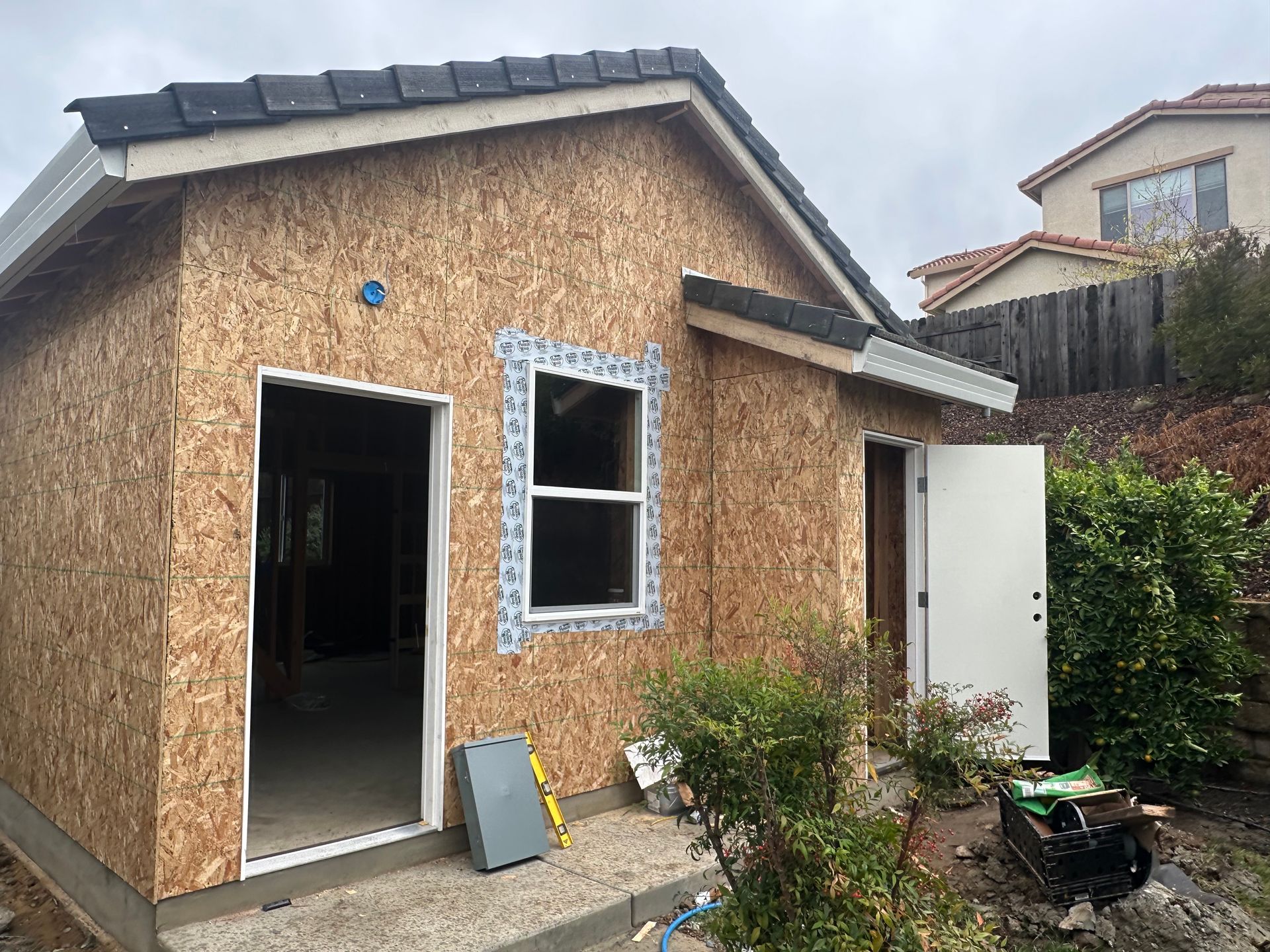 Small unfinished shed with OSB siding, open door, window, and garden tools beside it