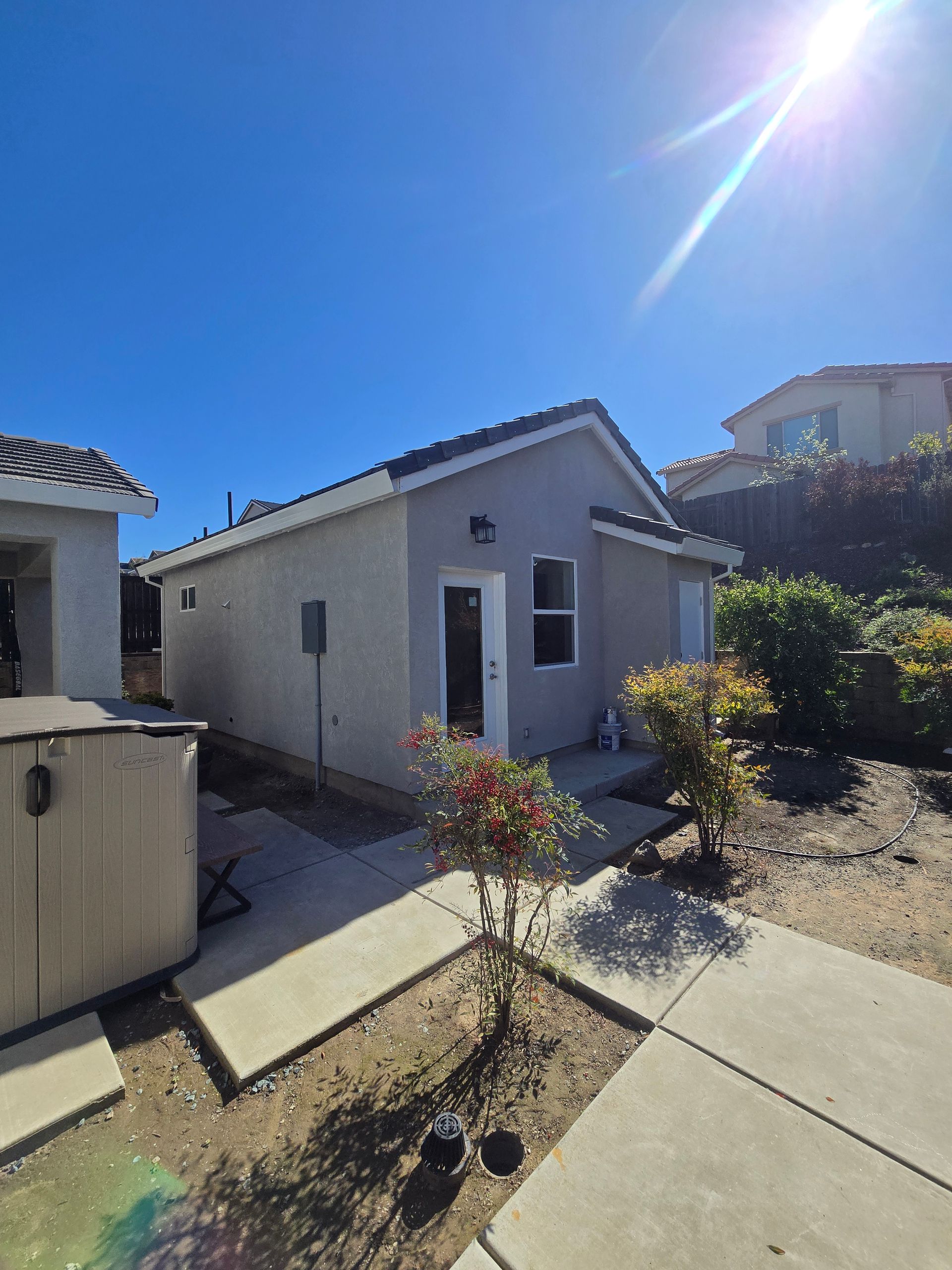 Small white house with a gray roof, concrete walkway, and shrubs under a bright blue sky.