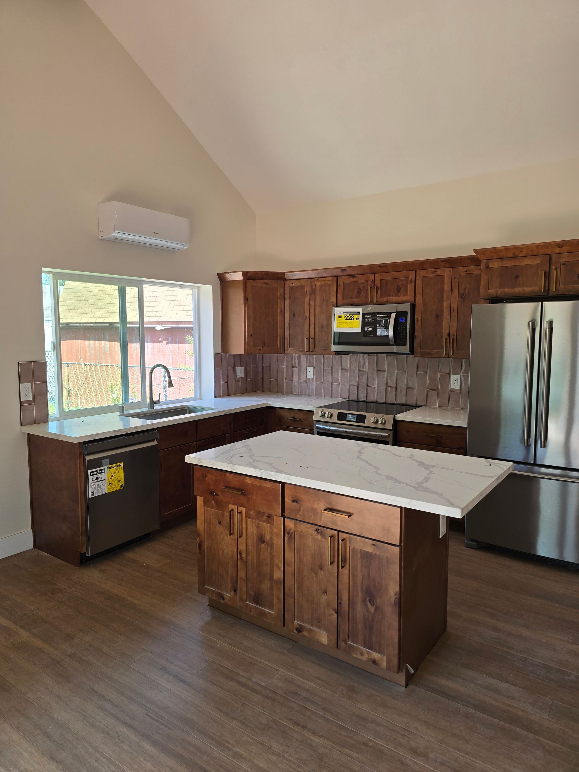 Empty kitchen with wood cabinets, white island, stainless fridge, and large window.
