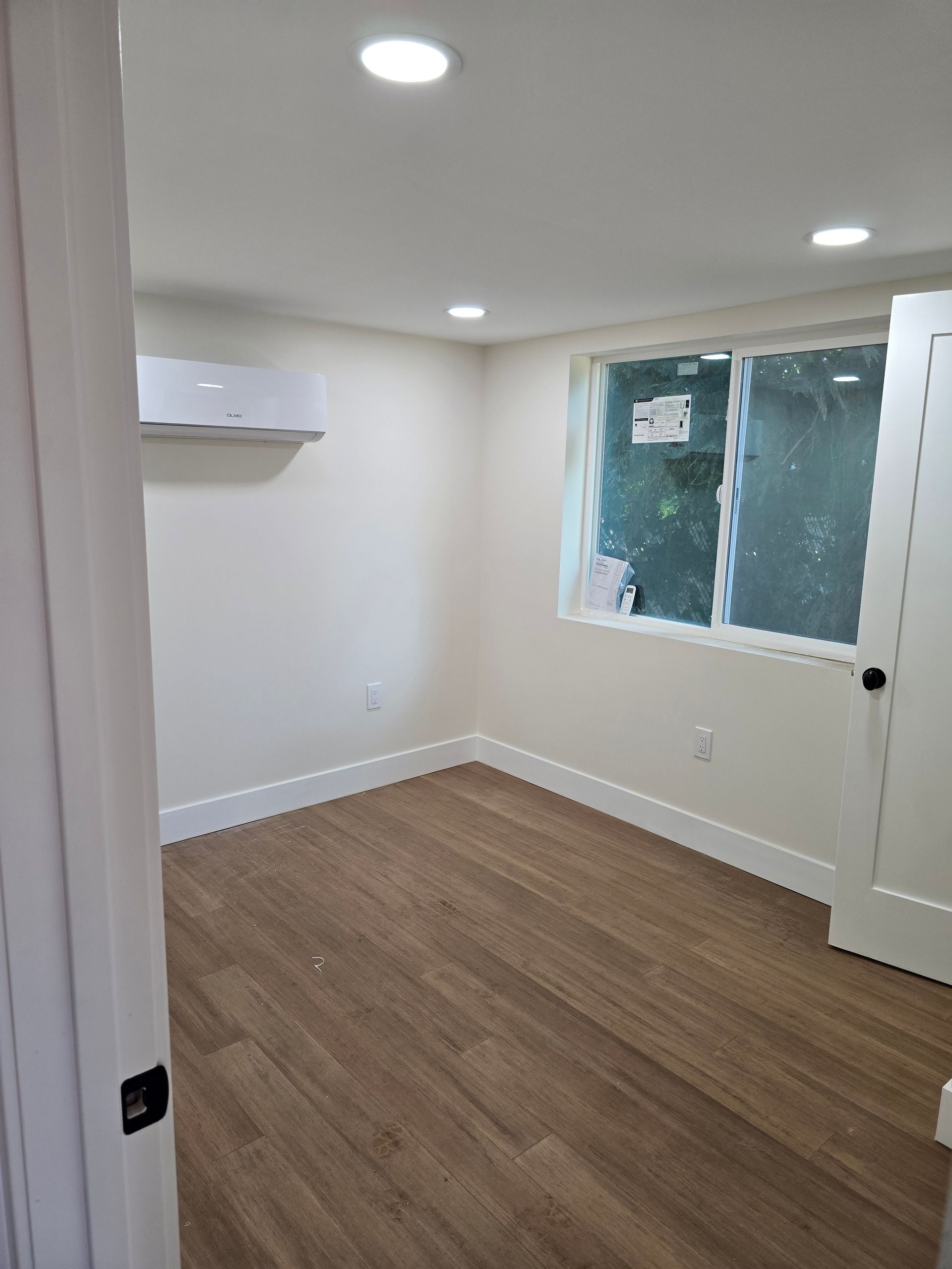 Empty small room with wood flooring, white walls, recessed lights, and a window on the right.