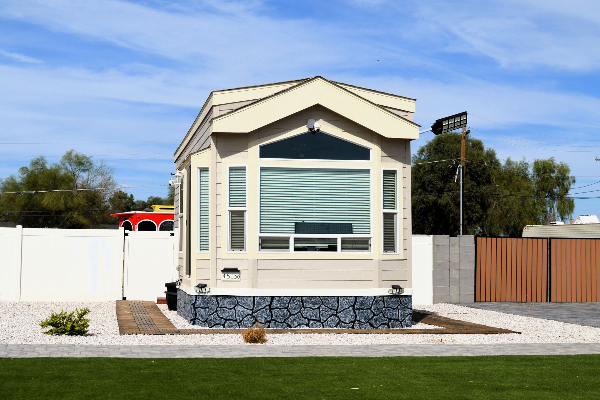 Small cream-colored modular home with large front window, gray stone base, and fenced yard under a blue sky