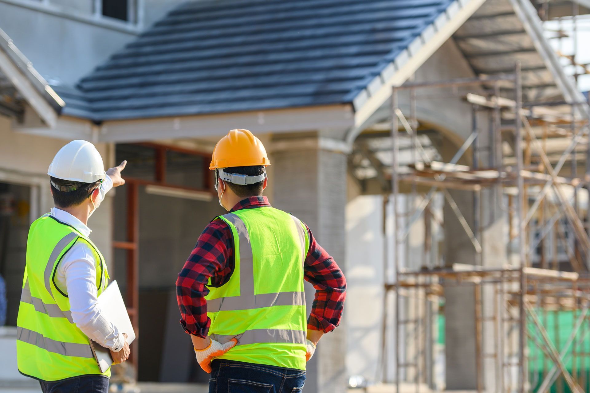 Two construction workers in safety vests and hard hats stand outside a house under construction.