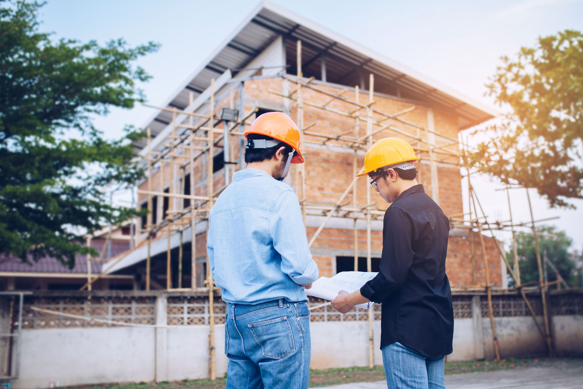 Two workers in hard hats review plans at a construction site with a building scaffold in the background.