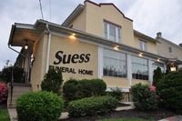 A beige-colored Suess Funeral Home building viewed from the exterior, featuring large windows and landscaped bushes.