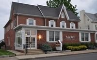A two-story brick building with white trim, serving as the Sadler Funeral Home, set beside a sidewalk and green lawn.