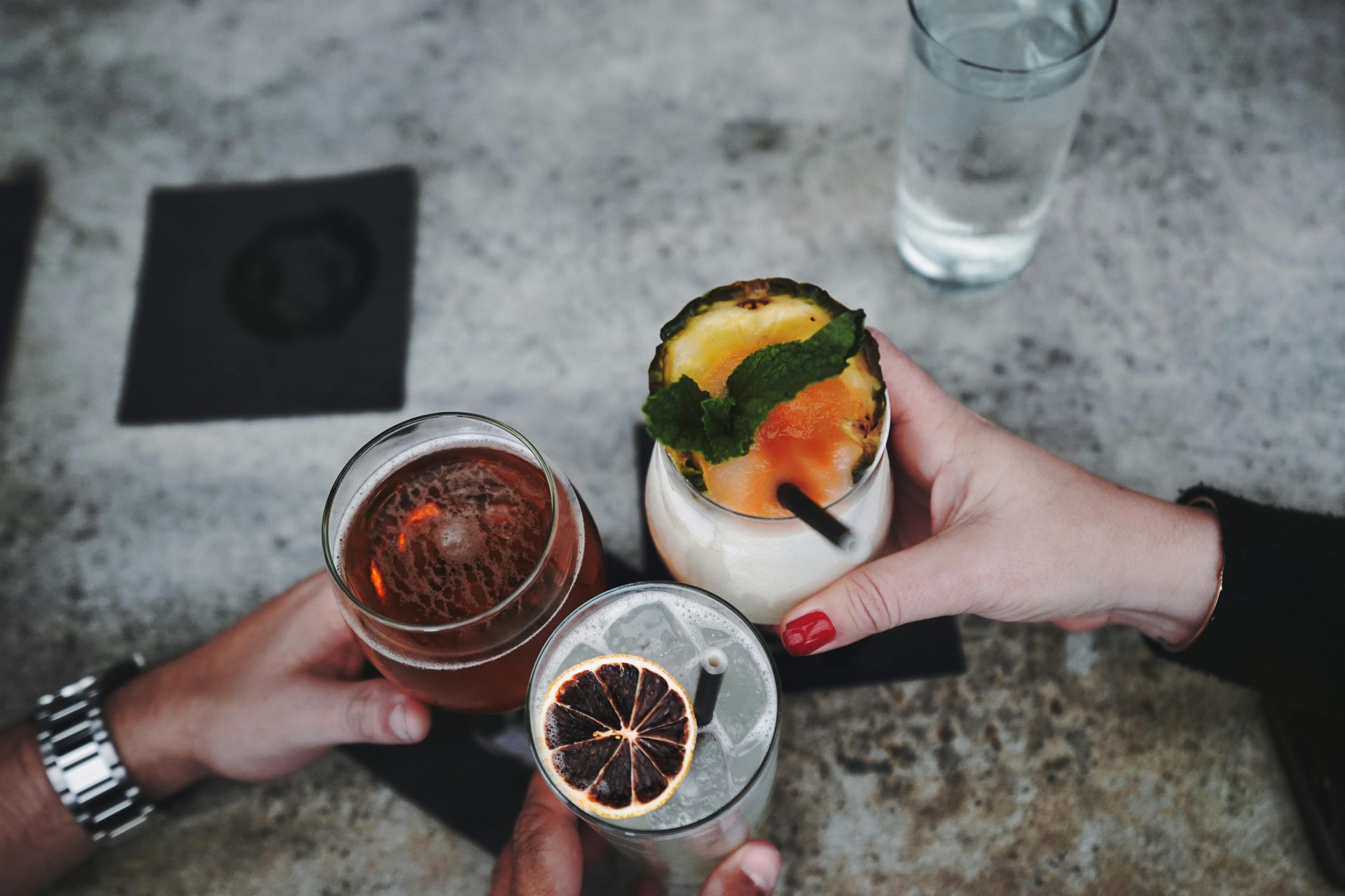 Hands toasting with three cocktails, a beer, and a glass of water on a gray concrete surface.