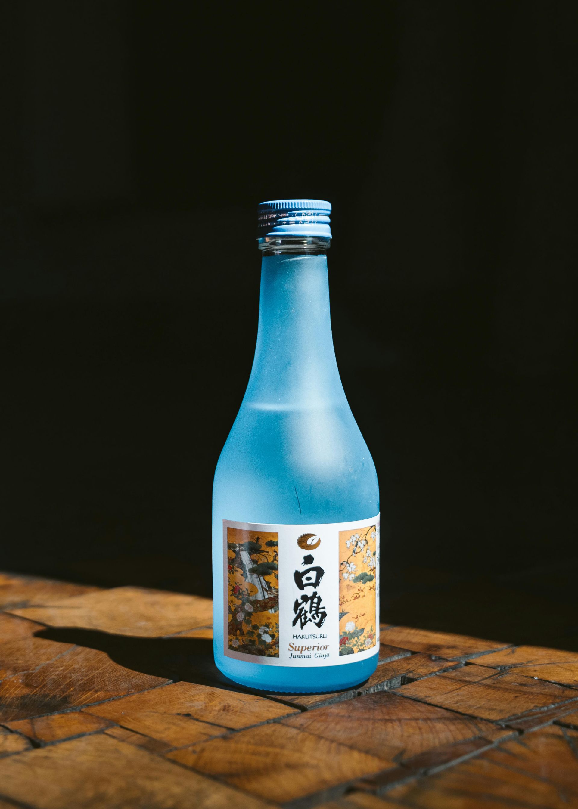 Blue sake bottle with a white and gold label, on a wooden surface with a dark background.