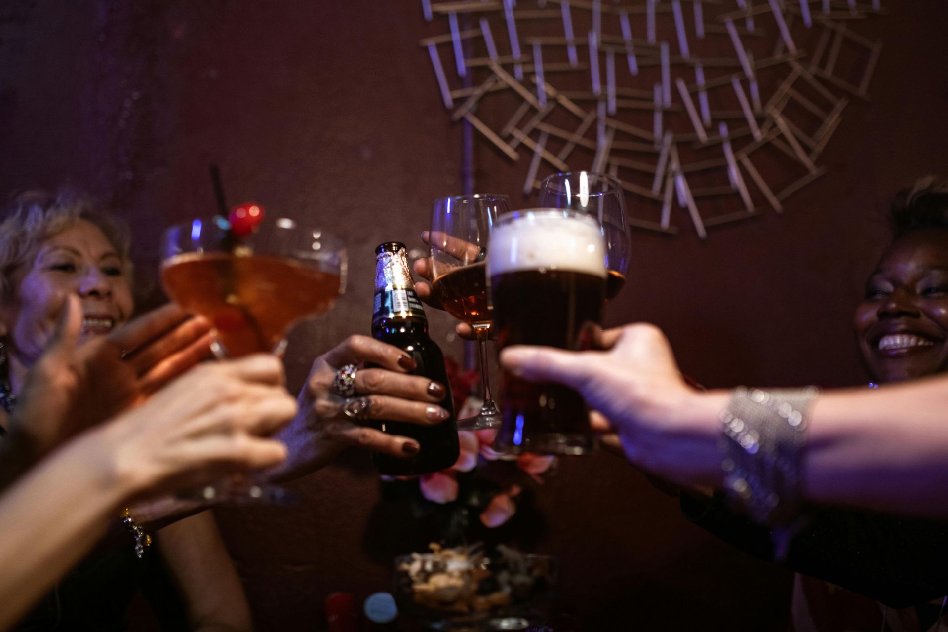 People toasting drinks in a dimly lit bar. Cheers with cocktails, beer, wine, and a bottle.