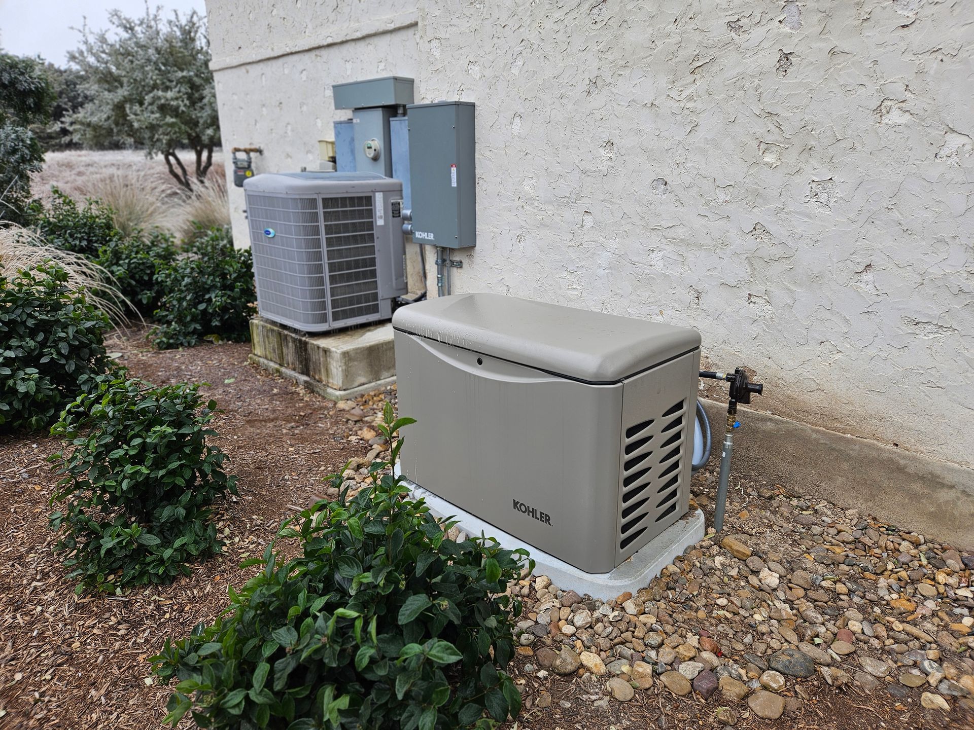 A Kohler generator, AC unit, and electrical panel against a stucco wall, surrounded by shrubs and rocks.