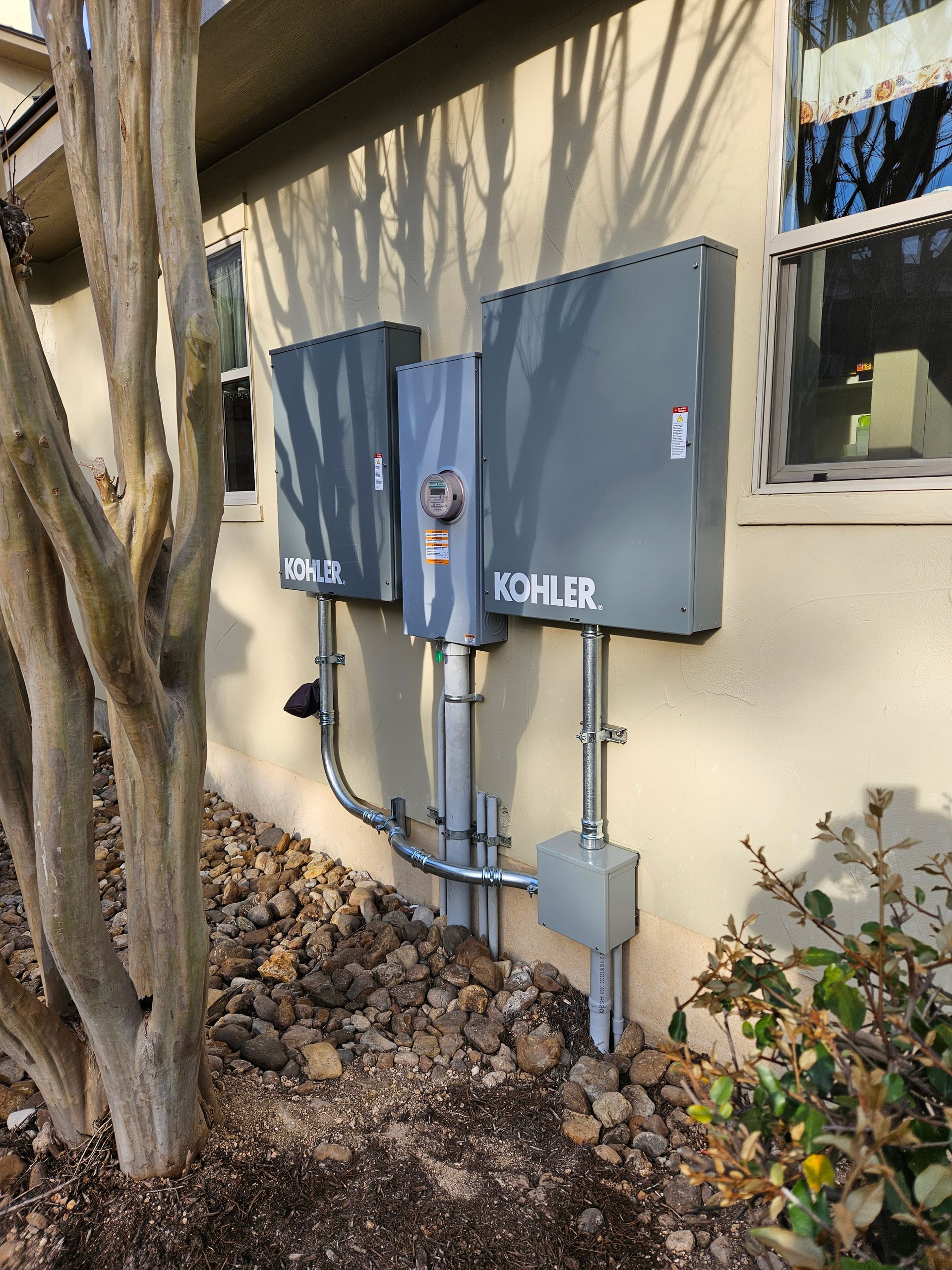 Three gray Kohler electrical boxes mounted on a beige house wall with conduit running down to a ground-level junction box.