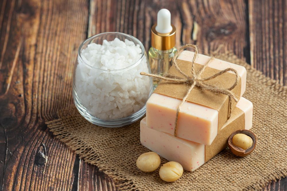 Soap bars, bath salts, oil dropper, and macadamia nuts on a wooden surface, suggesting skincare products.