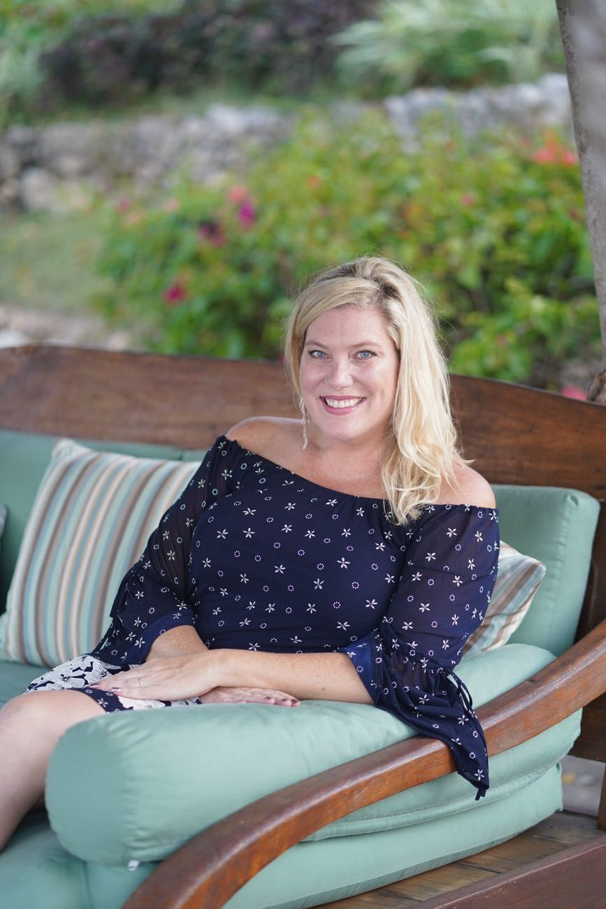 Woman with blonde hair, smiles in a navy off-the-shoulder dress, sitting on a green couch outdoors.