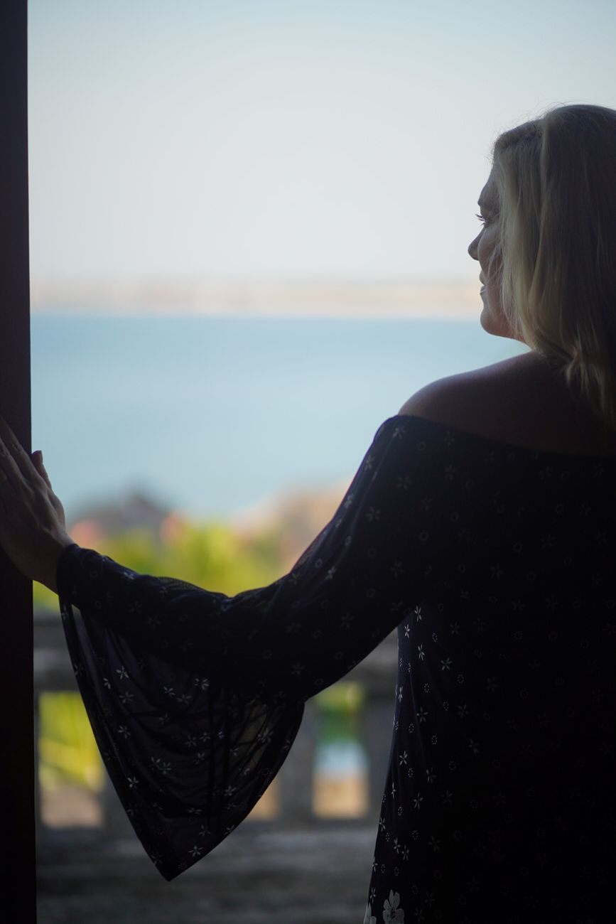 Woman in black dress looking out at ocean from a doorway.