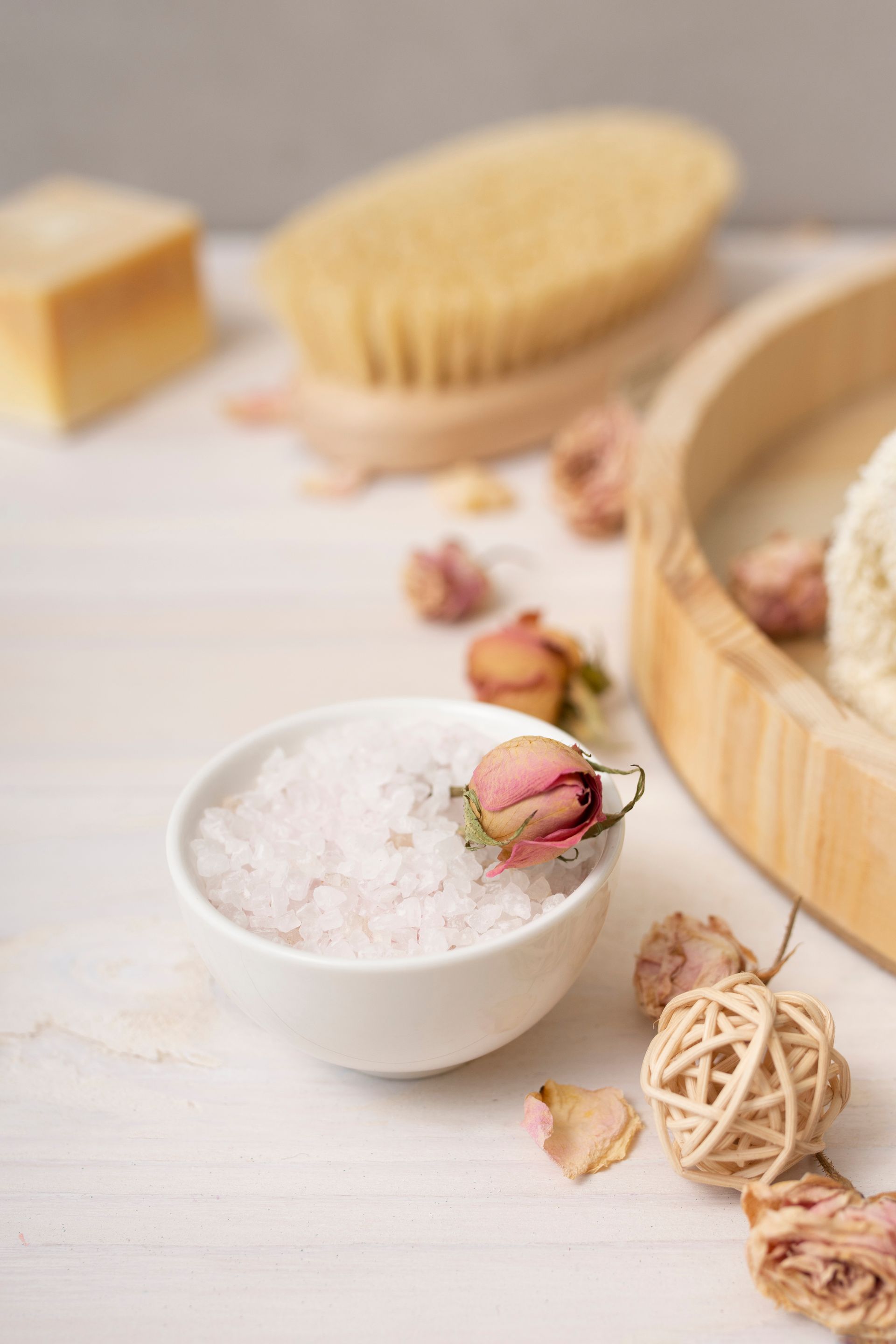 Spa scene with bath salts in a bowl, brush, soap, rosebuds, and wooden accents on a white surface.