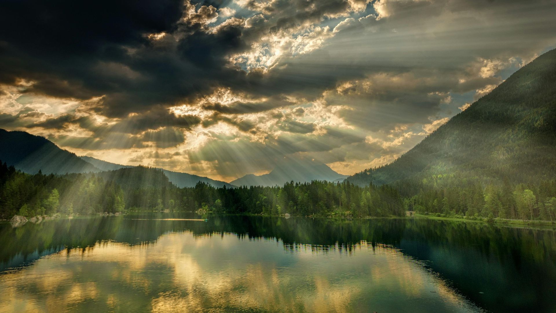 Sunlight beams through clouds over a calm lake reflecting mountains and trees.