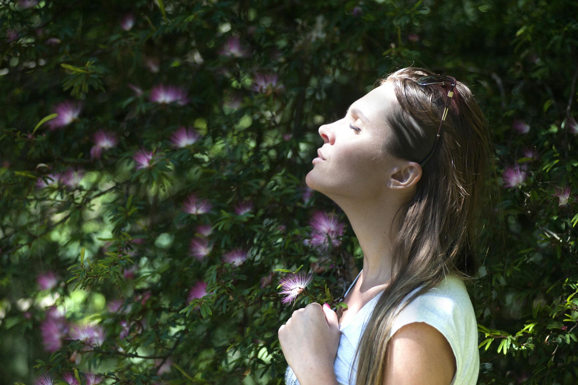 Woman with eyes closed, tilted head, basking in sunlight, in front of flowering bushes.