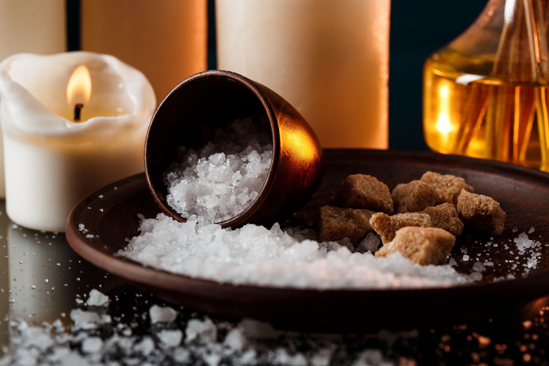 Candlelit scene: Salt spilling from a cup onto a plate with brown sugar cubes, candles in background.